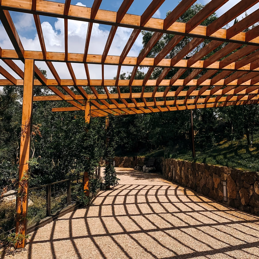 Timber pergola with closely spaced rafters casting a striped shadow pattern across a gravel path below